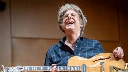 Pat Metheny laughs on stage as he holds a guitar in his lap.